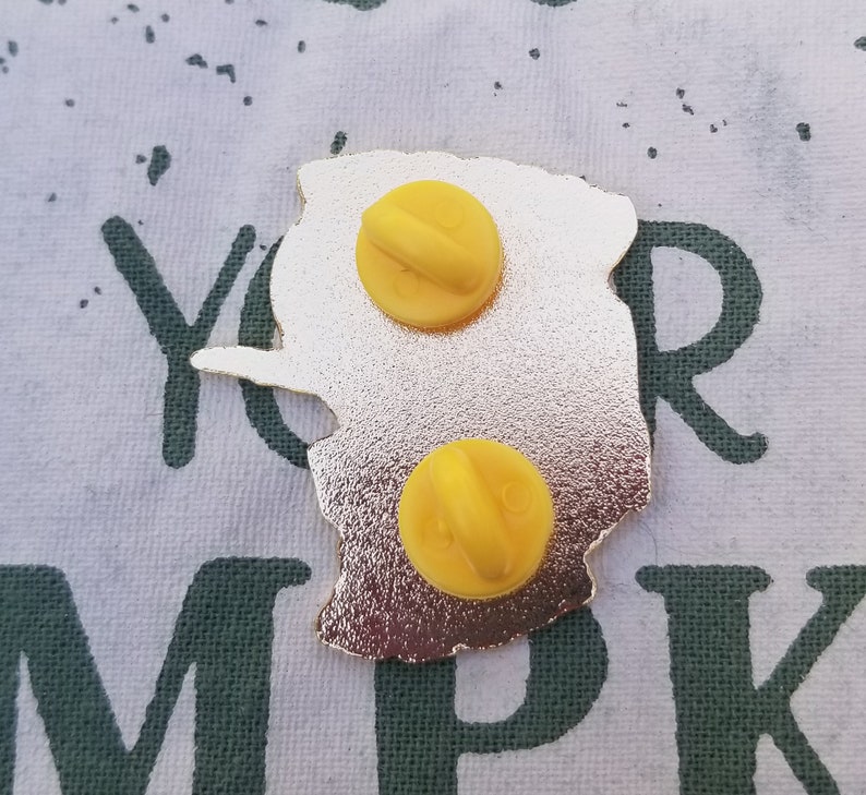 Pode incluir: O verso de um alfinete de metal dourado com um design amarelo. O alfinete tem dois fechos de metal amarelo na parte de tr&aacute;s.