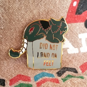 Puede incluir: Pin esmaltado con un gato zombi verde con detalles cosidos, descansando sobre una lápida. La lápida dice "DID NOT LAND ON FEET" en letras doradas, con un borde dorado alrededor del pin.