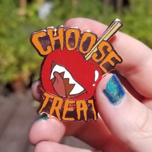 Pode incluir: Um broche de esmalte vermelho e laranja com as palavras "Choose Treat" e uma ma&ccedil;&atilde; vermelha de desenho animado com um sorriso de dentes e um pau que sai do topo.