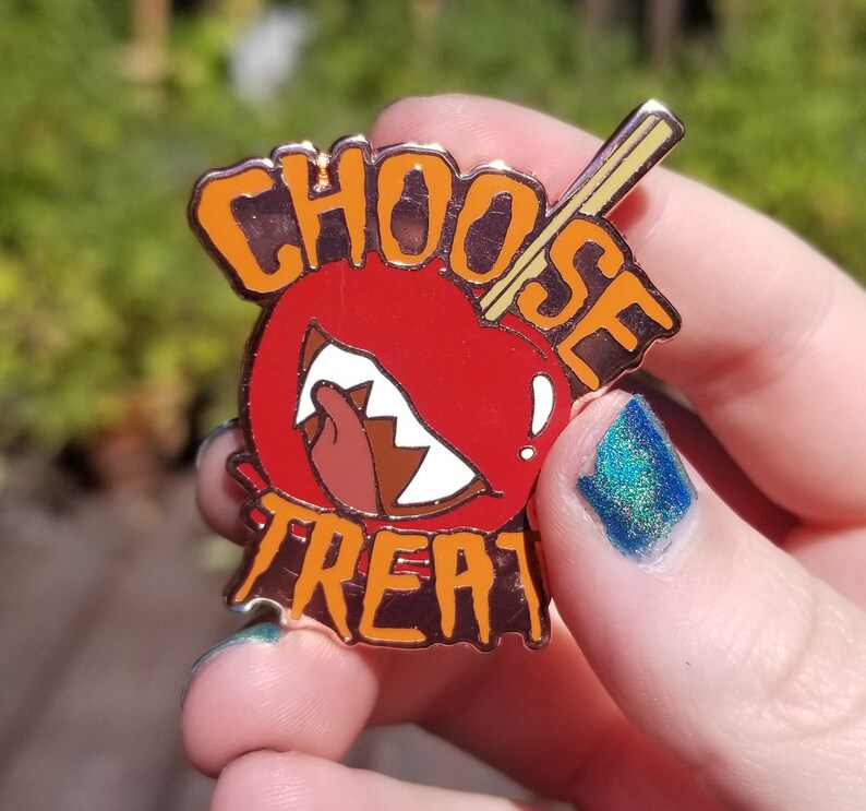 Pode incluir: Um broche de esmalte vermelho e laranja com as palavras "Choose Treat" e uma ma&ccedil;&atilde; vermelha de desenho animado com um sorriso de dentes e um pau que sai do topo.