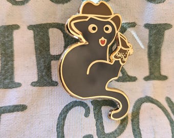 May include: A black cat-shaped enamel pin with a gold border, wearing a brown cowboy hat and holding a cupcake. The pin has a red mouth and two round eyes. The pin is on a white and green patterned background.