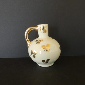 May include: A small, white ceramic pitcher with a round body and a gold handle. The pitcher is decorated with gold leaf clover designs. The handle and the rim of the pitcher are also gold. The pitcher is on a black surface.