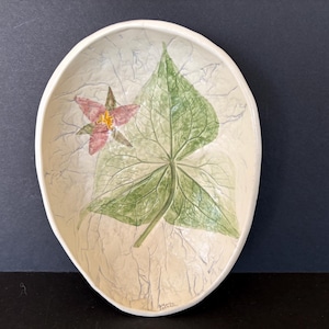 May include: Oval-shaped ceramic dish with a hand-painted floral design. The dish features a large green leaf and a pink flower with yellow accents. The background is a textured off-white color, and the dish has a slightly raised rim.