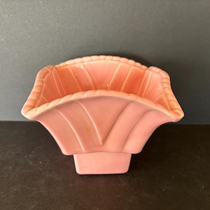 May include: A vintage, light pink ceramic basket-shaped planter. The planter has a scalloped rim and a fan-like design on the front. It sits on a rectangular base. The overall design is symmetrical and decorative.