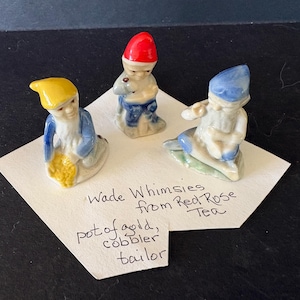 May include: Three Wade Whimsies figurines from Red Rose Tea. The figurines are of a pot of gold, a cobbler, and a tailor. The figurines are small, with the pot of gold figurine wearing a yellow hat and holding a yellow object.