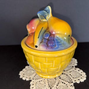 May include: A yellow ceramic fruit basket with a lid. The lid is shaped like a collection of colorful fruits, including bananas, grapes, and a peach. The basket has a woven texture and sits on a white lace doily.