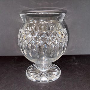 May include: A clear, cut-glass vase with a round body and a flared rim. The vase features a diamond pattern and sits on a pedestal base. The vase is approximately 6 inches tall.