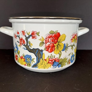 May include: White enamel pot with a floral design featuring red, yellow, and blue flowers and a brown tree branch. The pot has two white handles and a silver rim. A decorative kitchenware item.