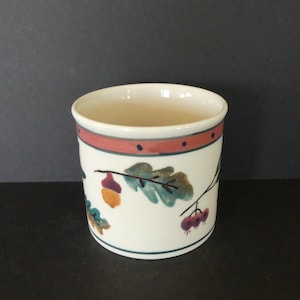 May include: A white ceramic tumbler with a decorative design of acorns, leaves, and berries. The rim features a red band with small black dots. The base has a thin blue line. The tumbler is cylindrical and has a smooth surface.
