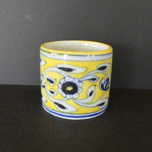 May include: A cylindrical ceramic cup with a yellow background and floral design. The design features blue and black flowers and white leaf-like shapes. The rim is white, and the base has a blue stripe. The cup is likely handmade.