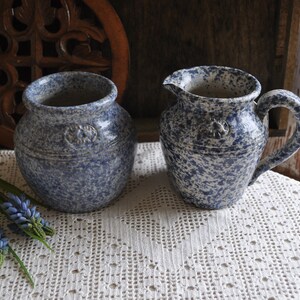 May include: Two blue and white speckled ceramic pieces. One is a small pitcher with a handle and spout, the other is a round vase. Both have a star-like emblem. They sit on a white lace cloth.