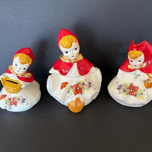 May include: Three ceramic figurines of girls dressed in red hooded cloaks and white dresses. Each girl is holding a basket and has a floral design on her dress.