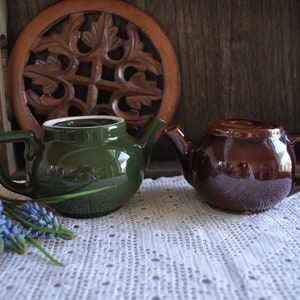 May include: Two ceramic teapots, one dark green and one brown, sit on a white lace doily. A small bouquet of blue flowers and a wooden trivet are in the background. The teapots have a classic design.