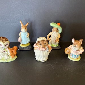 May include: A collection of ceramic figurines featuring animal characters. The figurines include an owl, a rabbit in a blue dress, a hedgehog, a rabbit in a hat, and a mouse. Each figurine is painted with detailed features and stands on a small base.