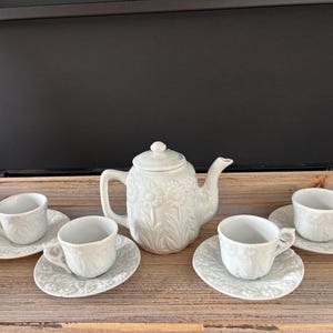 May include: A white ceramic tea set with floral detailing. The set includes a teapot with a lid, four teacups, and four saucers. The tea set is arranged on a wooden tray.