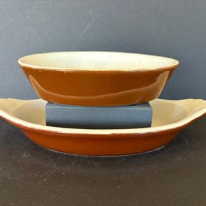May include: Two brown ceramic dishes, one oval and one round, stacked on a blue block. The dishes have a cream-colored interior and a brown exterior. The oval dish has decorative handles.