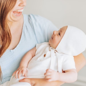 May include: A woman in a light blue dress holds a baby wearing a white onesie and a white bonnet. The baby is looking up at the woman and smiling.