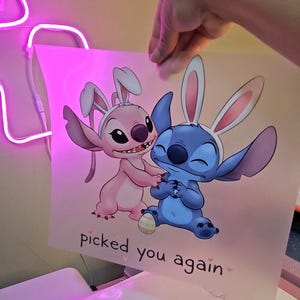 May include: A print featuring the characters Stitch and Angel from Lilo & Stitch, both wearing bunny ears. The pink and blue characters are on a light pink background with the text "picked you again".