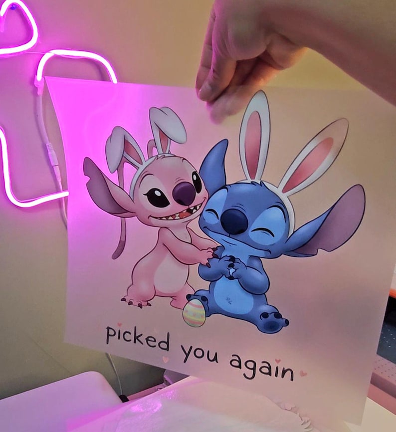 May include: A print featuring the characters Stitch and Angel from Lilo & Stitch, both wearing bunny ears. The pink and blue characters are on a light pink background with the text "picked you again".