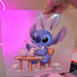 May include: A cartoon illustration of a blue alien character wearing bunny ears and a bow tie, painting Easter eggs. The character is seated at a small wooden table with paint and eggs. The background is a soft pink.