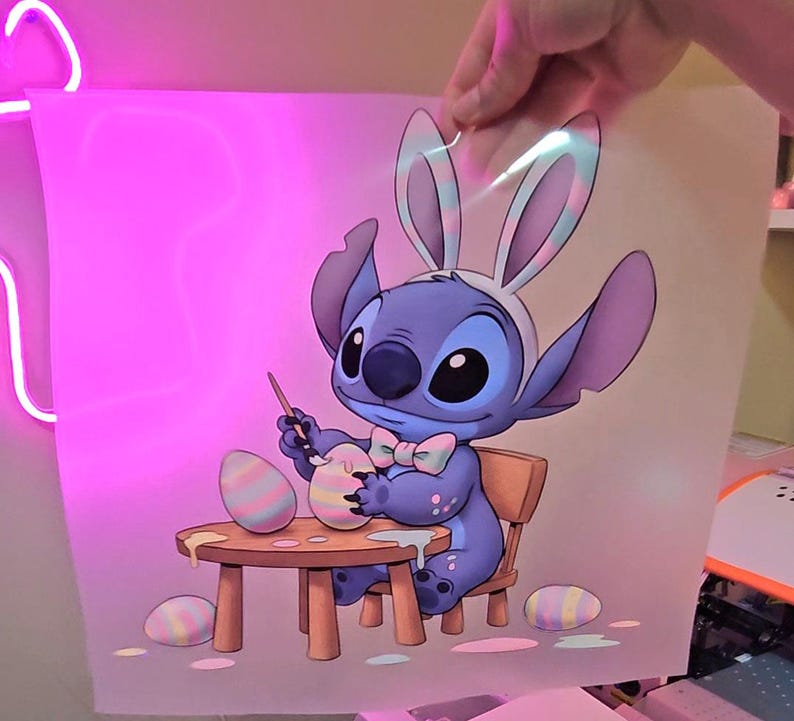 May include: A cartoon illustration of a blue alien character wearing bunny ears and a bow tie, painting Easter eggs. The character is seated at a small wooden table with paint and eggs. The background is a soft pink.