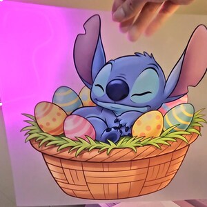 May include: A cartoon illustration of Stitch from Lilo & Stitch, nestled in a woven basket filled with colorful Easter eggs and green grass. Stitch is blue with large ears and is sleeping peacefully.