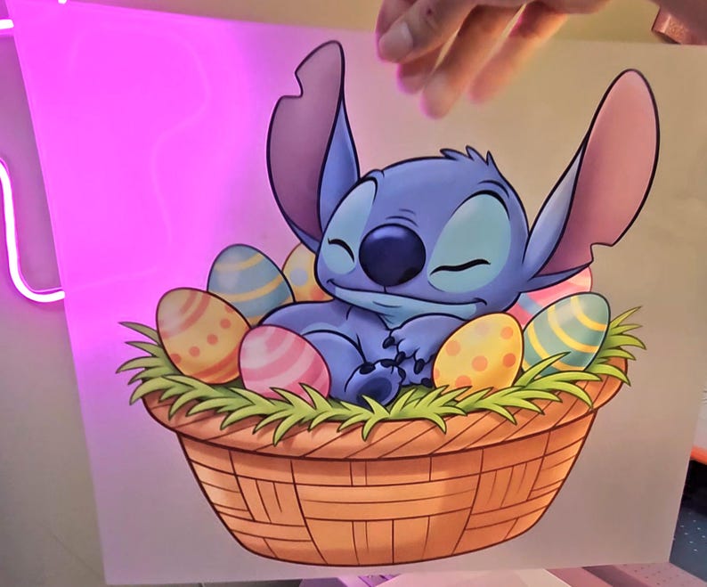 May include: A cartoon illustration of Stitch from Lilo & Stitch, nestled in a woven basket filled with colorful Easter eggs and green grass. Stitch is blue with large ears and is sleeping peacefully.