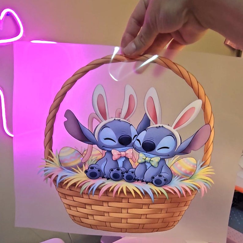 May include: A cartoon image of two blue aliens wearing bunny ears and bow ties, sitting in a woven basket filled with colorful Easter eggs and straw. The basket has a brown handle and is set against a pink background.