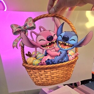 May include: A cartoon image of Stitch and Angel from Lilo & Stitch in a woven basket. Angel is pink, and Stitch is blue, both smiling. The basket contains Easter eggs and a bow. A hand is holding the image.