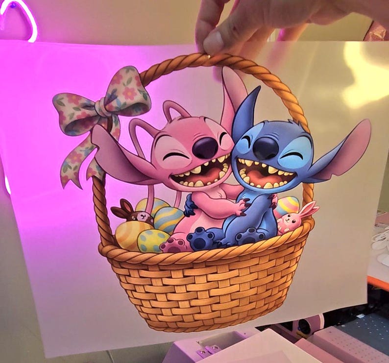 May include: A cartoon image of Stitch and Angel from Lilo & Stitch in a woven basket. Angel is pink, and Stitch is blue, both smiling. The basket contains Easter eggs and a bow. A hand is holding the image.
