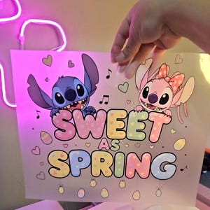 May include: A pink and white graphic featuring the characters Stitch and Angel from Lilo & Stitch. The text "Sweet as Spring" is displayed in pastel colors, with musical notes, hearts, and Easter eggs. The image is held by a hand.