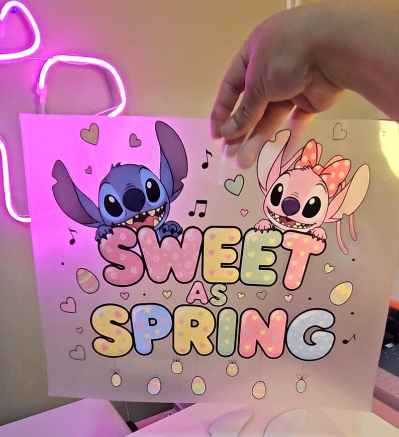 May include: A pink and white graphic featuring the characters Stitch and Angel from Lilo & Stitch. The text "Sweet as Spring" is displayed in pastel colors, with musical notes, hearts, and Easter eggs. The image is held by a hand.