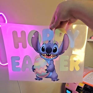 May include: A cartoon image of Stitch from Lilo & Stitch holding a pastel Easter egg. The image has the words "Happy Easter" in pastel colors. Stitch is wearing a flower crown. The image is held up by a hand.