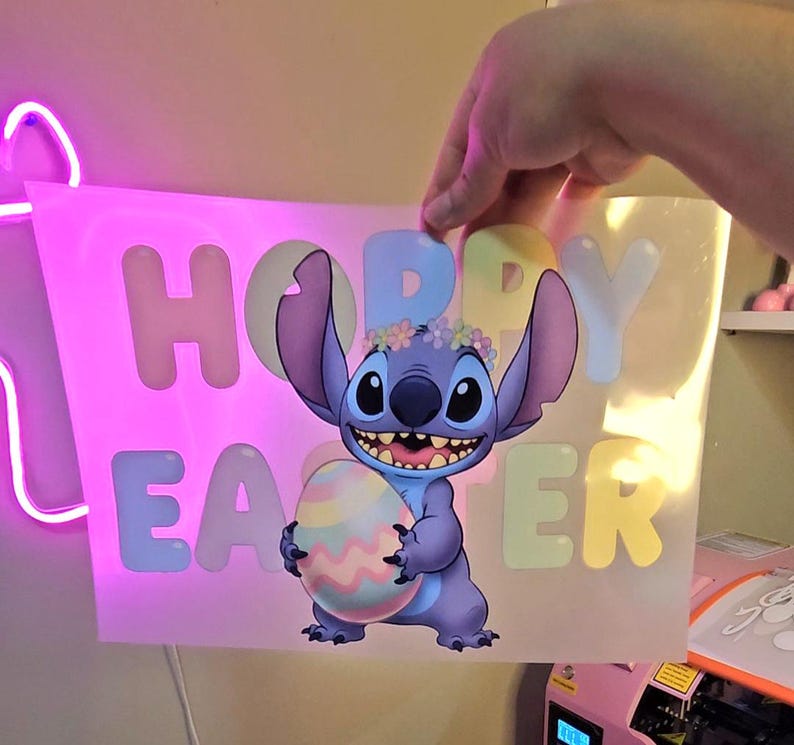 May include: A cartoon image of Stitch from Lilo & Stitch holding a pastel Easter egg. The image has the words "Happy Easter" in pastel colors. Stitch is wearing a flower crown. The image is held up by a hand.