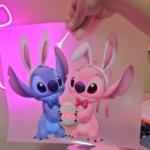May include: A cartoon illustration of Stitch and Angel, both wearing bunny ears and holding an Easter egg. Stitch is blue, Angel is pink, and the egg is decorated with pastel colors. The image is held up by a hand.