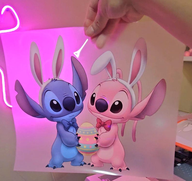 May include: A cartoon illustration of Stitch and Angel, both wearing bunny ears and holding an Easter egg. Stitch is blue, Angel is pink, and the egg is decorated with pastel colors. The image is held up by a hand.