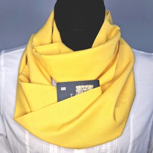 May include: A bright yellow infinity scarf featuring a hidden zippered pocket. A black rectangular card with a white graphic design is tucked inside the pocket, which is integrated into the fabric folds of the scarf worn around the neck.