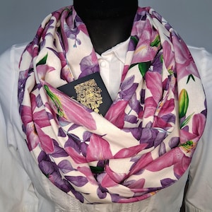 Könnte beinhalten: Ein floraler Rundschal mit cremefarbenem Hintergrund, verziert mit großen rosa und lila Lilienblüten und grünen Blättern. Ein schwarzer Reisepass mit goldenen Details ist in einer Tasche zu sehen. Der Schal ist über einem weißen Hemd drapiert.