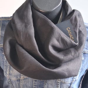 May include: A dark charcoal gray infinity scarf draped in loose folds, featuring a hidden pocket containing a dark blue passport with the word CANADA printed in gold lettering, worn over a blue denim jacket on a mannequin torso.