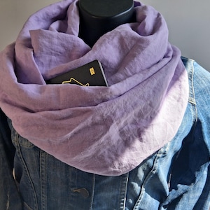 May include: A lavender infinity scarf draped around a mannequin neck, featuring a hidden zippered pocket containing a black passport with a gold airplane icon. The scarf is worn over a blue denim jacket with visible metal buttons and stitching.
