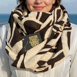 May include: An infinity scarf featuring a bold, abstract pattern of dark brown shapes on a cream-colored background. A small, rectangular black tag with a gold-colored, intricate, embossed design is attached to the front of the fabric.
