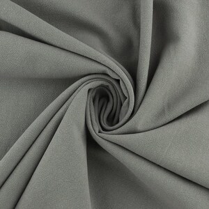 Op de afbeelding: Een close-up van een grijze stofstaal, strak gedraaid om de textuur en de drapering te laten zien. De stof heeft een licht ruwe, matte afwerking en de plooien cre&euml;ren schaduwen die de diepte en dimensie benadrukken. De kleur is een gedempt, neutraal grijs.