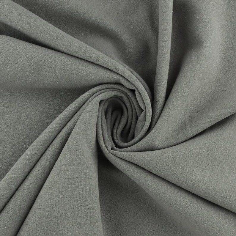 Op de afbeelding: Een close-up van een grijze stofstaal, strak gedraaid om de textuur en de drapering te laten zien. De stof heeft een licht ruwe, matte afwerking en de plooien cre&euml;ren schaduwen die de diepte en dimensie benadrukken. De kleur is een gedempt, neutraal grijs.
