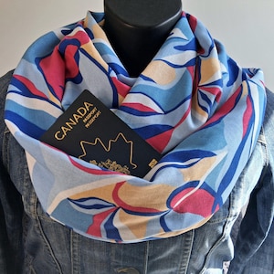 May include: A light blue infinity scarf featuring an abstract pattern of pink, dark blue, and beige shapes. A dark blue Canada passport with gold lettering and a maple leaf emblem is tucked into the folds of the fabric, worn over a denim jacket.