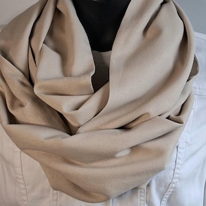 May include: A solid light beige infinity scarf draped in multiple loose folds around the neck. The fabric appears smooth and matte, creating a continuous loop design that rests against a white denim jacket worn underneath the accessory.