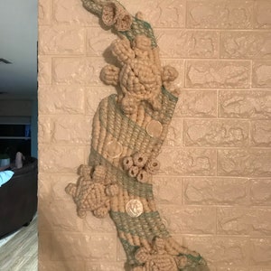 May include: A wall hanging made of woven yarn and rope in shades of beige and green. The hanging features a series of crocheted sea turtles and seashells.