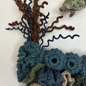 May include: A crocheted wall hanging featuring a brown and blue tree with green, blue, and brown coral-like shapes. A green crocheted turtle hangs from the top of the tree.