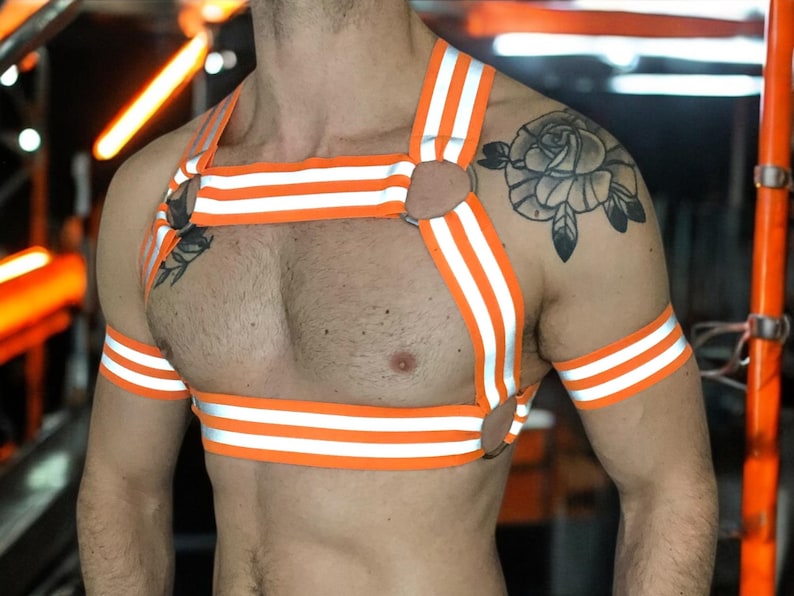 Made-to-measure men’s elastic chest harness in high-visibility neon orange with reflective detailing, glowing under blacklight. The design features double shoulder and chest straps joined by circular O-rings, with optional matching jockstrap and thigh bands—ideal for clubwear, festivals, or fetish fashion.
