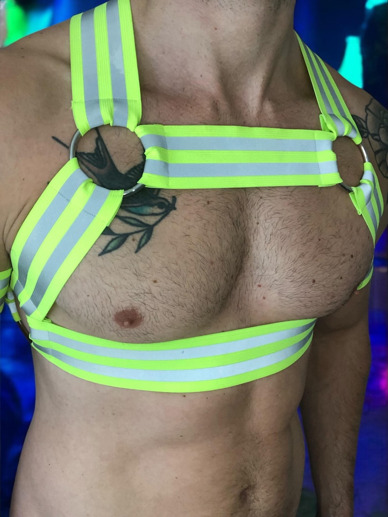 Made-to-measure men’s elastic chest harness in high-visibility neon yellow with reflective detailing, glowing under blacklight. The design features double shoulder and chest straps joined by circular O-rings, with optional matching jockstrap and thigh bands—ideal for clubwear, festivals, or fetish fashion.