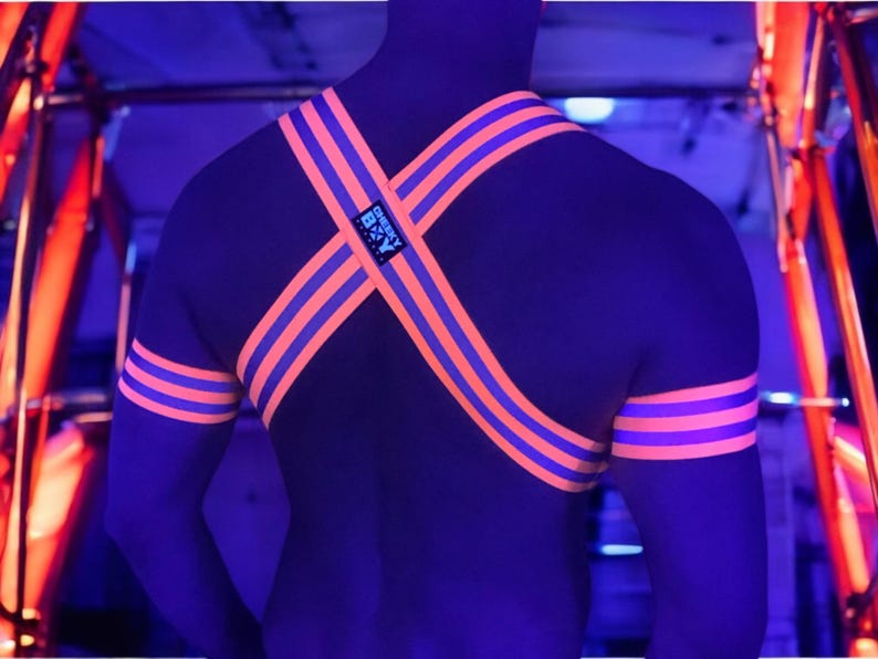 May include: A black harness with orange and purple stripes, worn over the shoulders and arms. The harness has a small tag with the text "Cheeky B&Y". The image is taken in a dark environment with neon lighting.